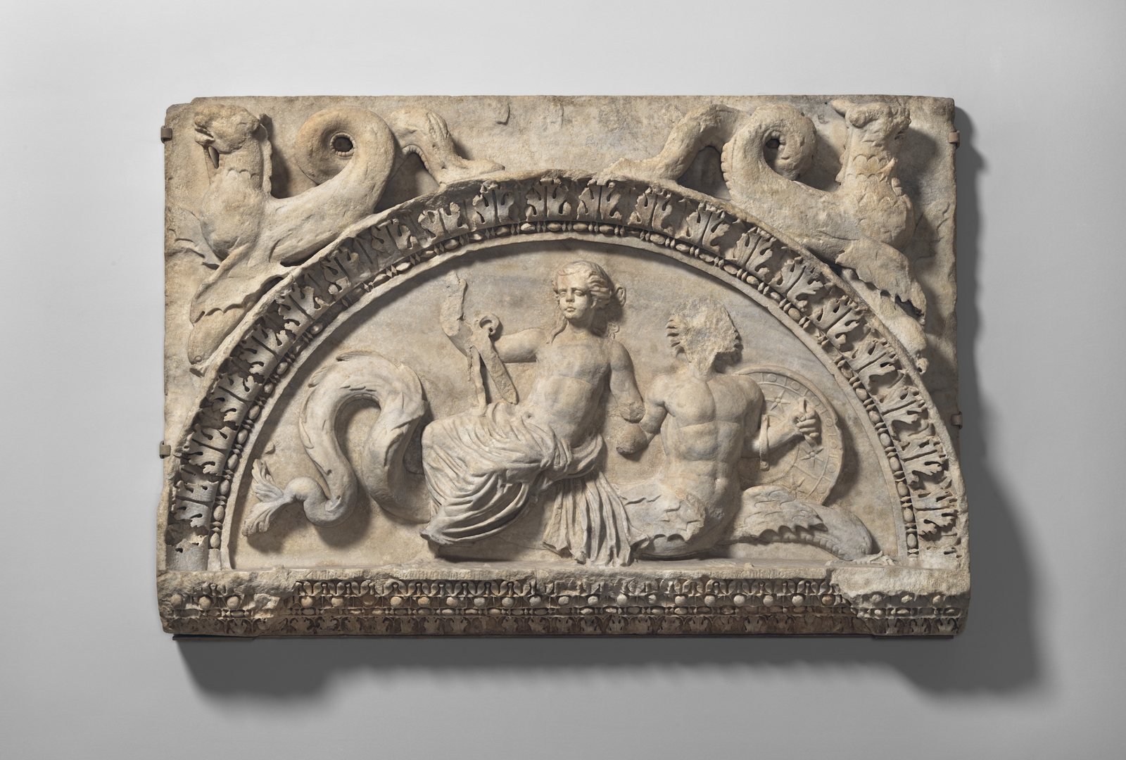 Roman marble lunette, early 2nd century AD. A Nereid sits on the tail of a Triton, a sea god with a fish's tail; she holds a sword and he carries a shield, the armor the Nereids brought to Achilles in the Iliad.